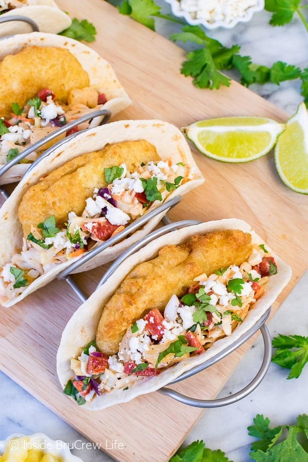 Two flour tortillas filled with crispy fish, slaw, cheese, and cilantro on a wooden cutting board with lime slices