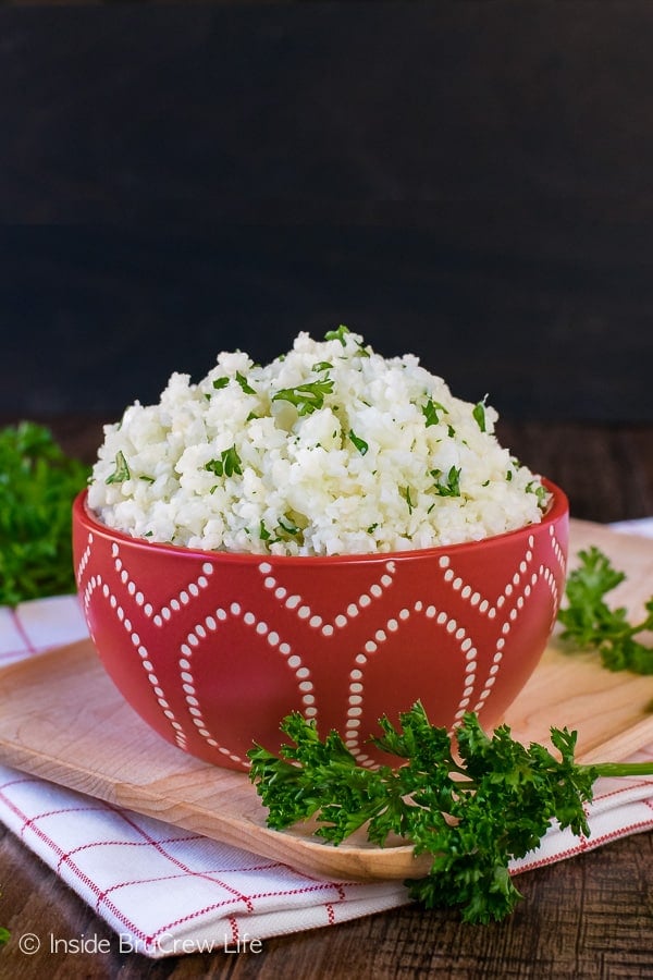 A wooden cutting board with a red bowl filled with homemade cauliflower rice on it.