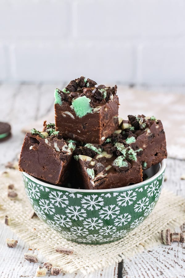 Multiple pieces of chocolate fudge loaded with green mint cookies and mint chips in a green bowl with white snowflakes.