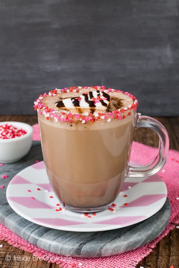 A glass cup of Dark Chocolate Latte with a rim of pink and red heart sprinkles sitting on a pink and white plate