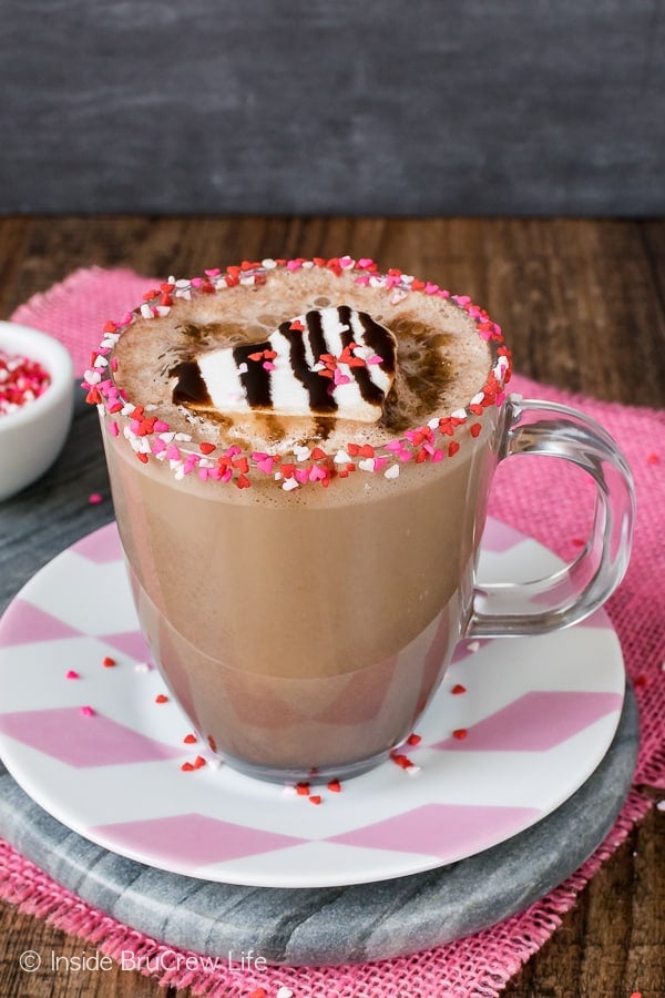 A picture of a glass of Dark Chocolate Latte on white and pink plate. The glass has a rim of pink and red heart sprinkles and a heart marshmallow is in the drink.