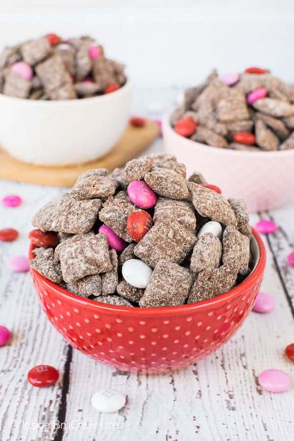 A close up picture of a red bowl filled with M&M's Brownie Batter Muddy Buddies and two bowls in the background.