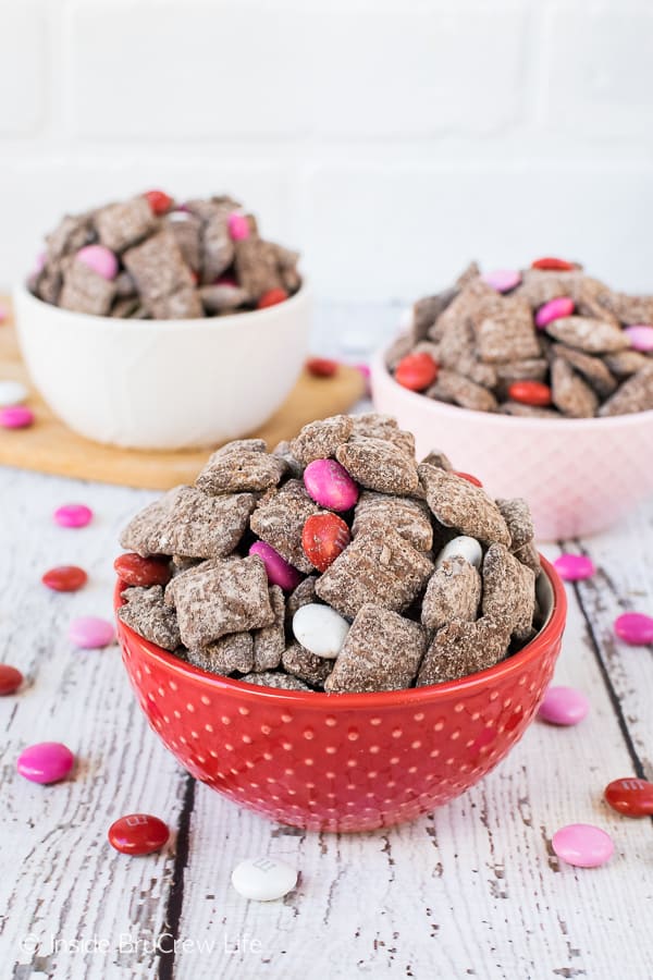 A picture of a red bowl with M&M's Brownie Batter Muddy Buddies in it with a white and pink bowl full of snack mix in the background