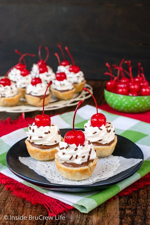 Three mini chocolate cheesecake bites topped with whipped cream and cherries on a black plate.