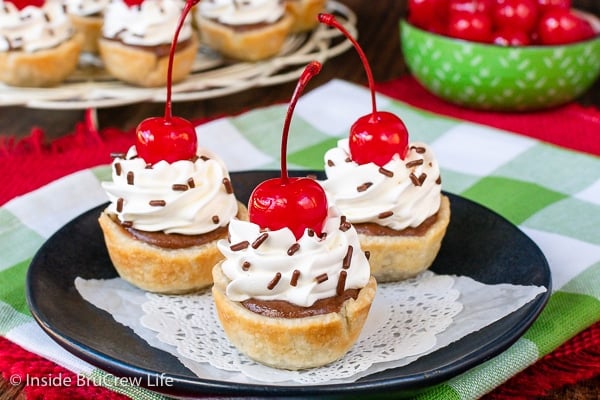 A black plate with three mini chocolate cheesecake bites topped with whipped cream, cherries, and sprinkles on it.