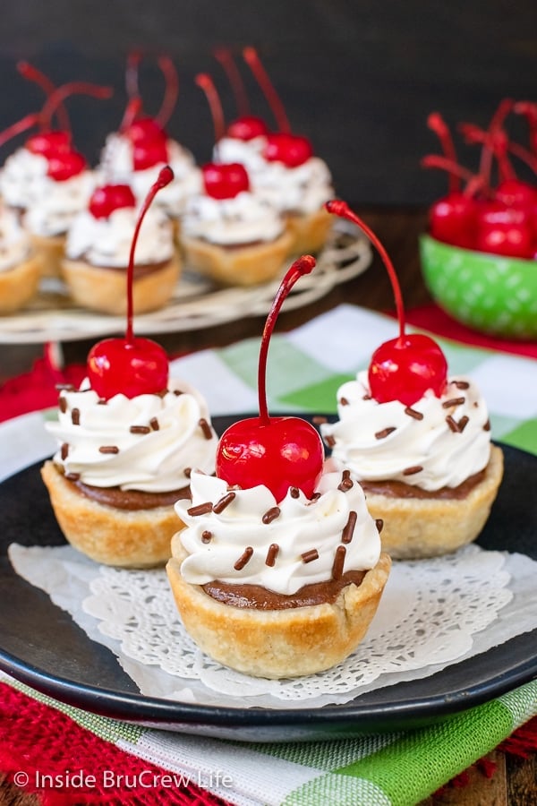 Three mini chocolate cheesecake bites topped with whipped cream and cherries on a black plate.