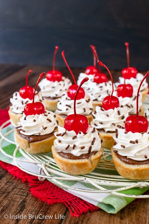 A white wire tray with a bunch of mini chocolate cheesecake bites topped with whipped cream and cherries on it.