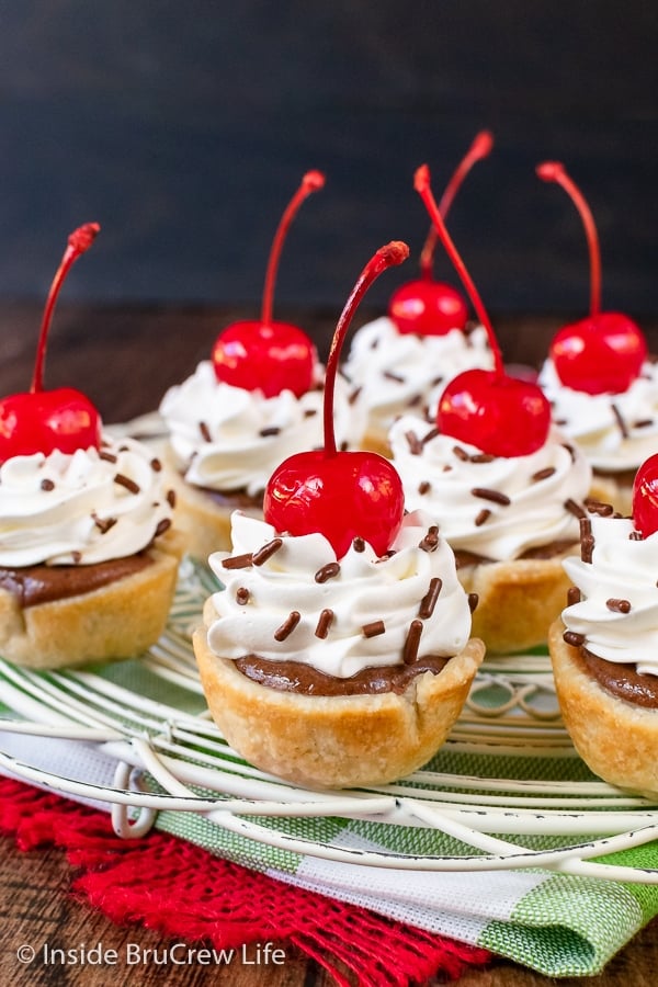 Mini chocolate pie bites topped with whipped cream and cherries on a white wire rack.