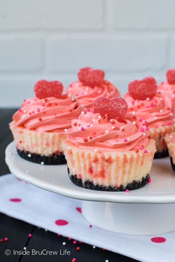 A close up picture of a white cake stand on a white napkin with Red Hot White Chocolate Cheesecakes on top. Sprinkles and jelly hearts add a fun flair to the pink mousse topping.