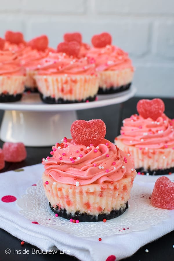 Two Red Hot White Chocolate Cheesecakes on a white napkin with a white cake stand behind with more cheesecakes on it.