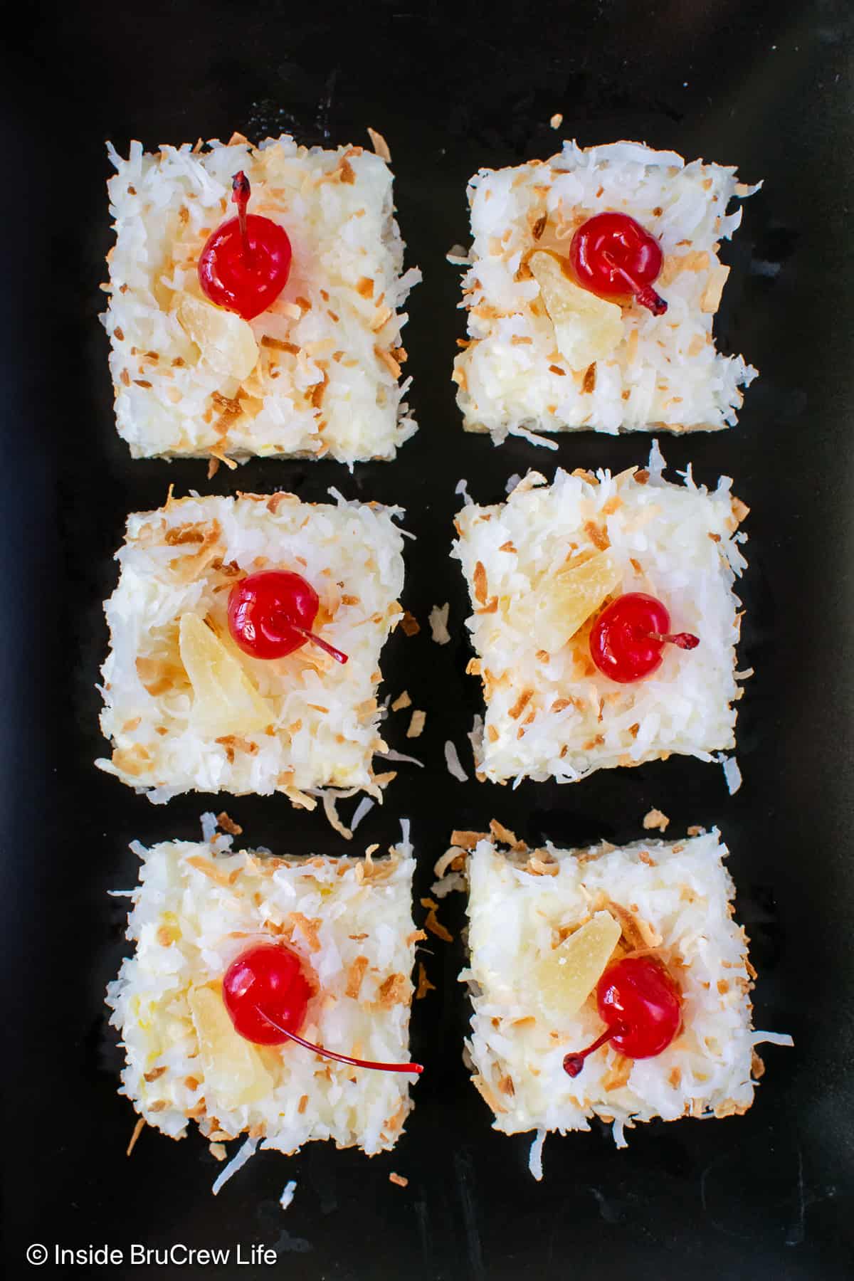 Six pina colada bars with toasted coconut and cherries on a black plate.