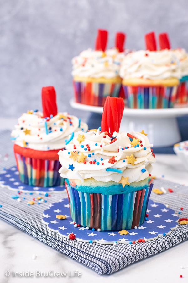 Two firecracker cupcakes in red white and blue liners topped with vanilla frosting, Twizzlers, and sprinkles.
