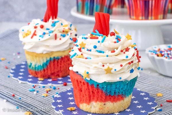 Two red white and blue cupcakes decorated with vanilla frosting, sprinkles, and Twizzlers on a blue towel.