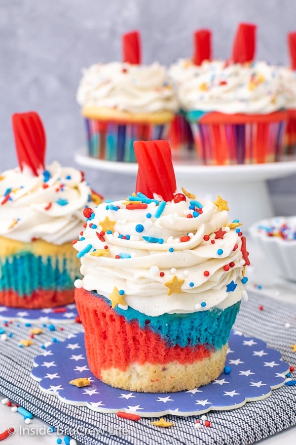 Two red white and blue firecracker cupcakes with vanilla frosting, sprinkles, and twizzlers on blue coasters. 