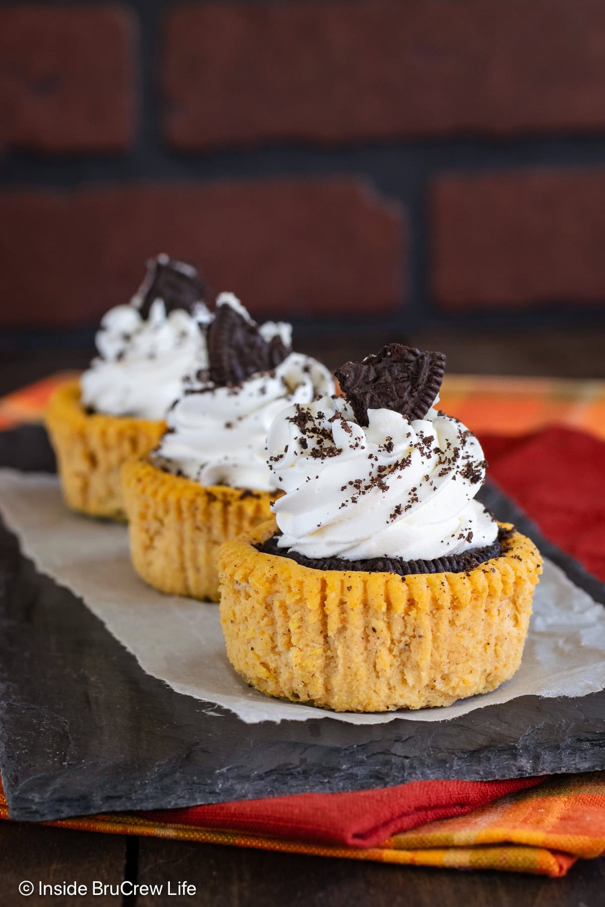 Three pumpkin Oreo cheesecakes topped with whipped cream on a black board.