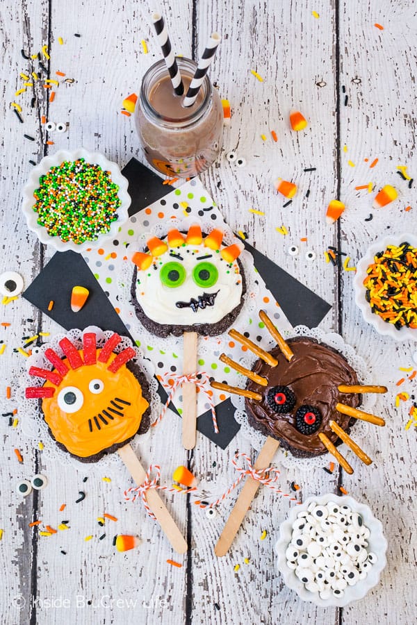 White background with monster sugar cookies on sticks on it and sprinkles and candy all around them.