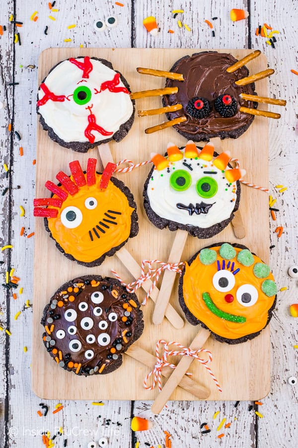 Overhead picture of a wooden cutting board with six decorated monster sugar cookies on popsicle sticks on it.