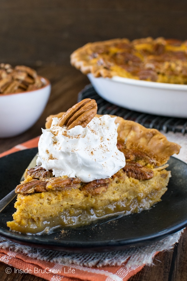 A slice of pecan pie cheesecake topped with Cool Whip and a pecan on a black plate.