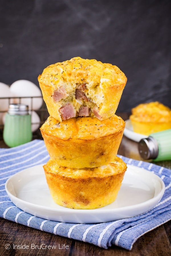 Three egg muffin cups stacked on a white plate with a bite out of the top one showing the ham and cheese inside