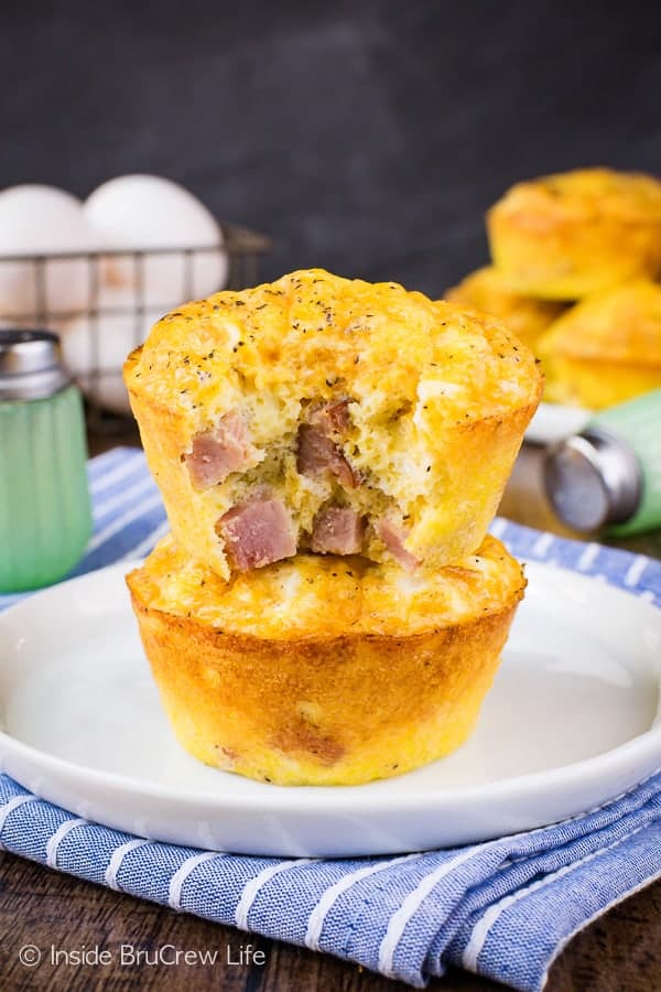 Two egg muffin cups stacked on a white plate with bite out of one showing the ham and cheese inside
