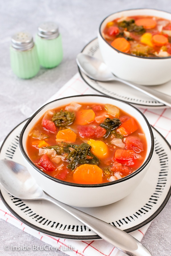 Two white bowls on white plates filled with a colorful vegetable soup.