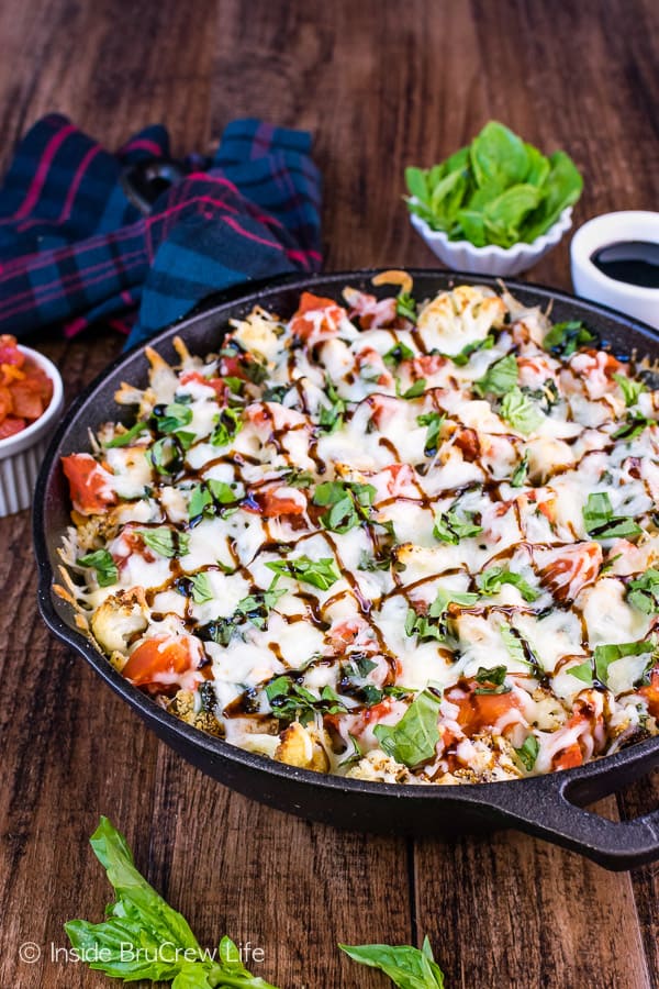 A cast iron skillet on a brown board filled with cheesy cauliflower nachos.