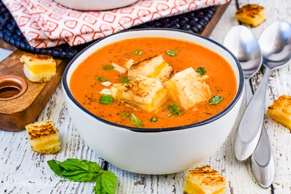 A white bowl on a white board filled with homemade creamy tomato soup and topped with grilled cheese bites
