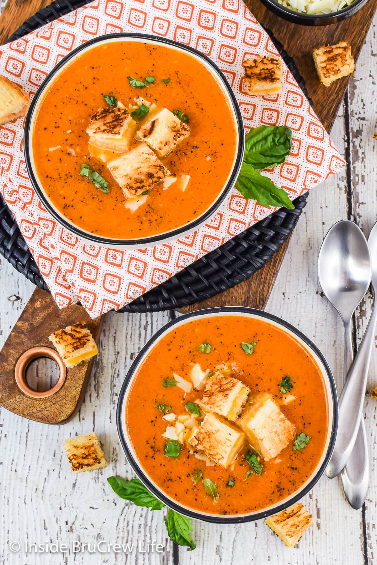 Two bowls of homemade tomato soup topped with basil and grilled cheese cubes.