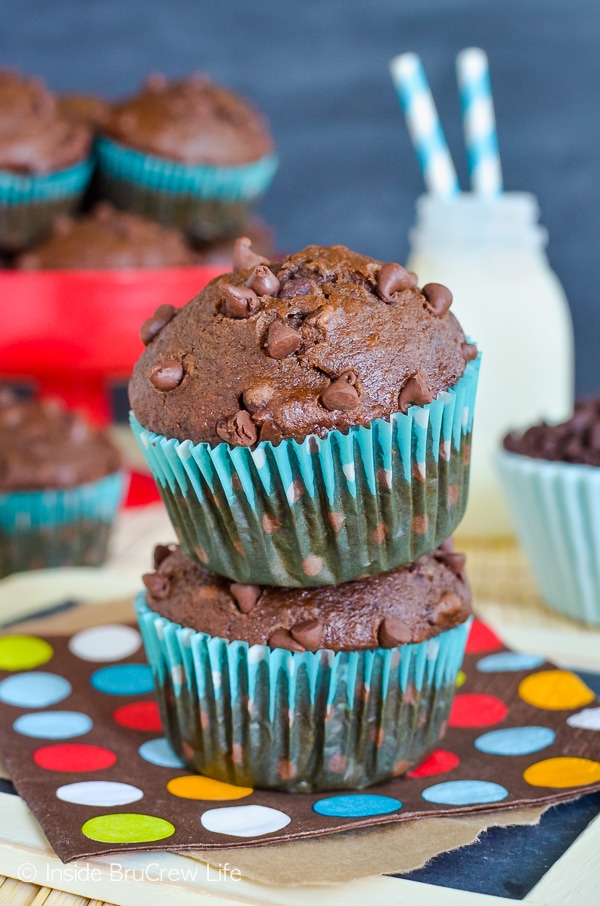 Two chocolate banana muffins stacked on top of each other on a colorful napkin. More muffins and a jug of milk are behind them.