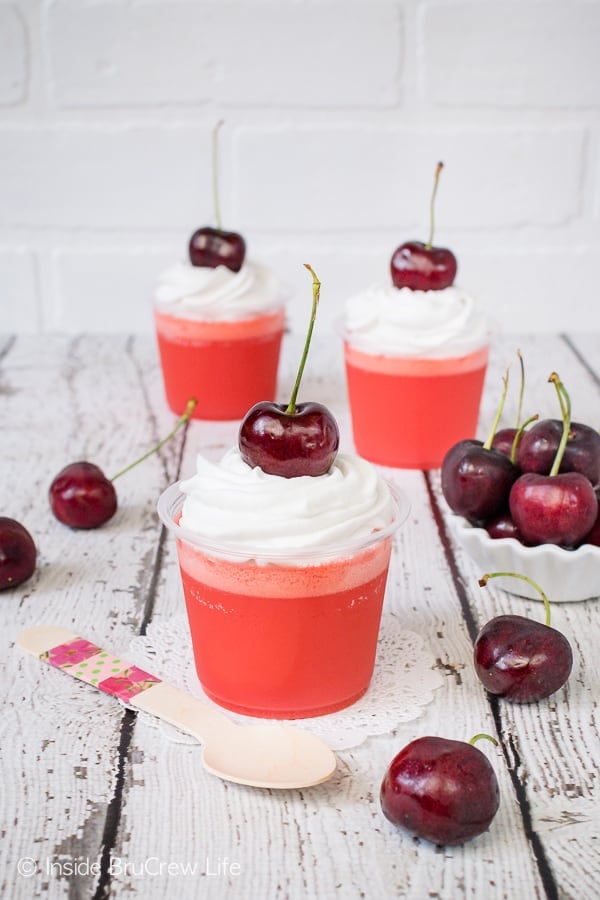 A white background with clear cups filled with cherry jello parfaits and topped with whipped cream and a cherry