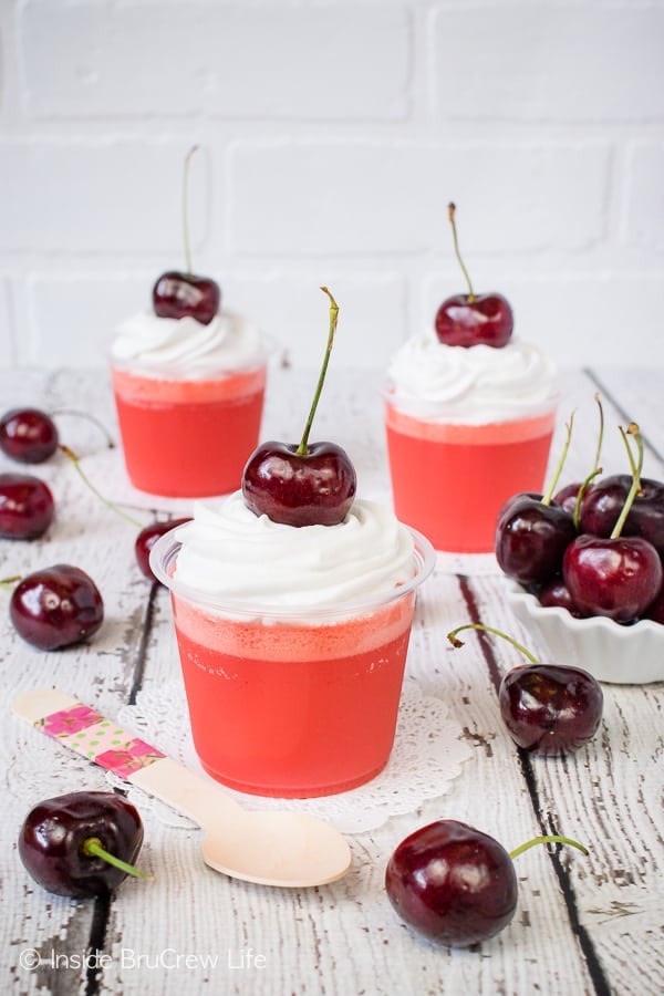 Clear parfait cups on a white background filled with layers of cherry jello and topped with whipped cream and a cherry