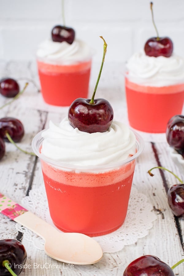 Three clear cups on a white background filled with low carb cherry jello parfaits and topped with whipped cream and a cherry