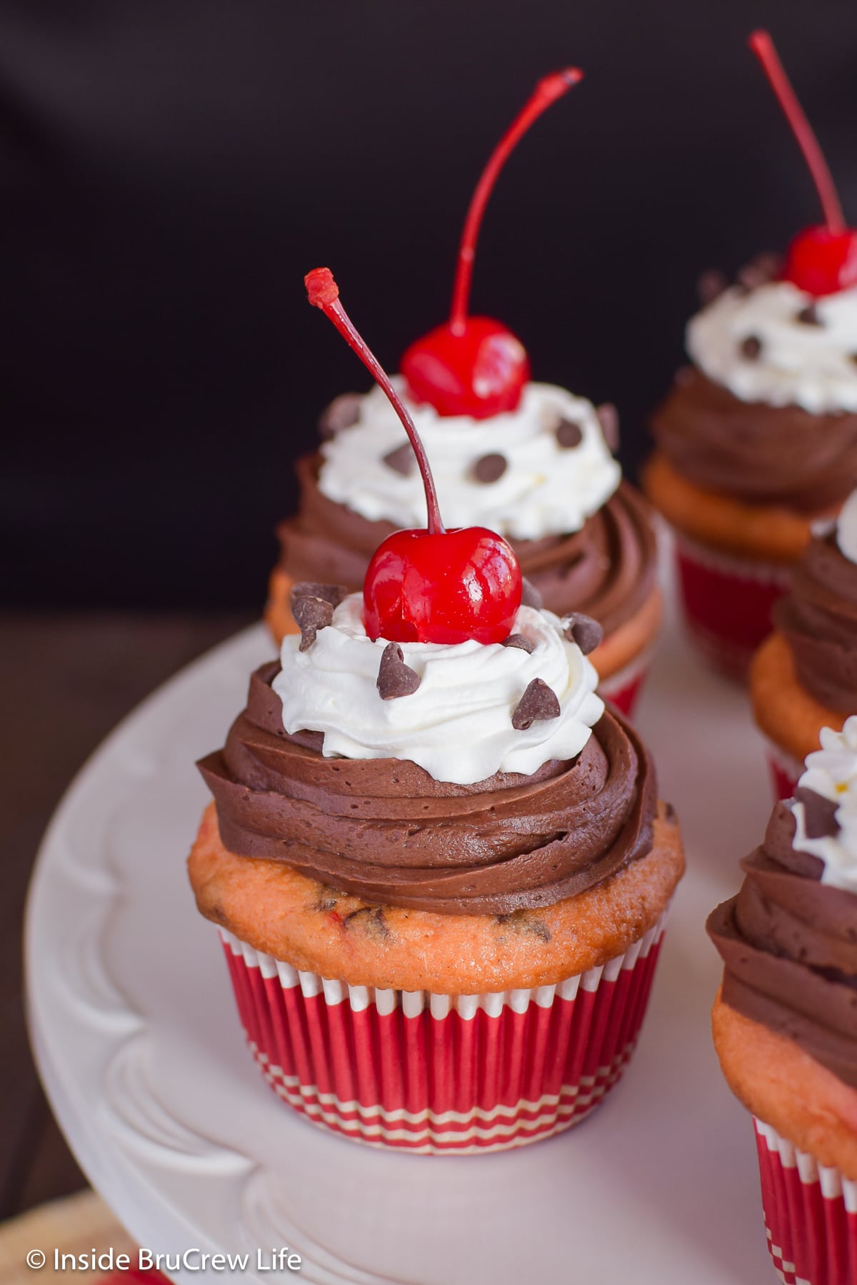 Cherry cupcakes topped with frosting and cherries on a cake plate.