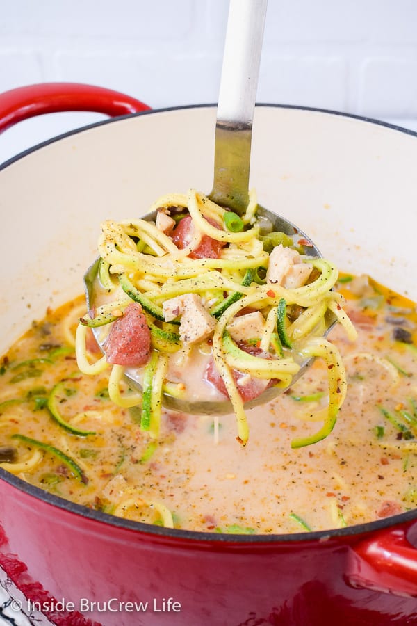 A ladle lifting out a big scoop of chicken zoodle soup from a red pot.