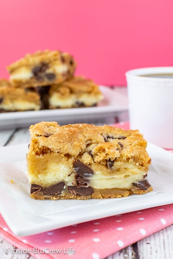 A white plate with a square of chocolate chip cheesecake bars on it with more bars behind it.