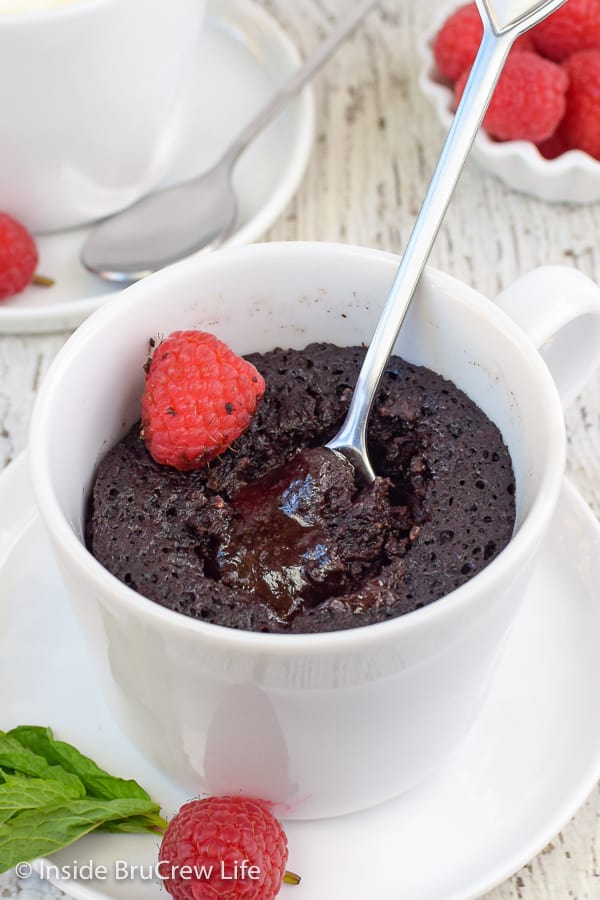 A white cup with a cooked chocolate lava cake in it and spoon showing the gooey center.