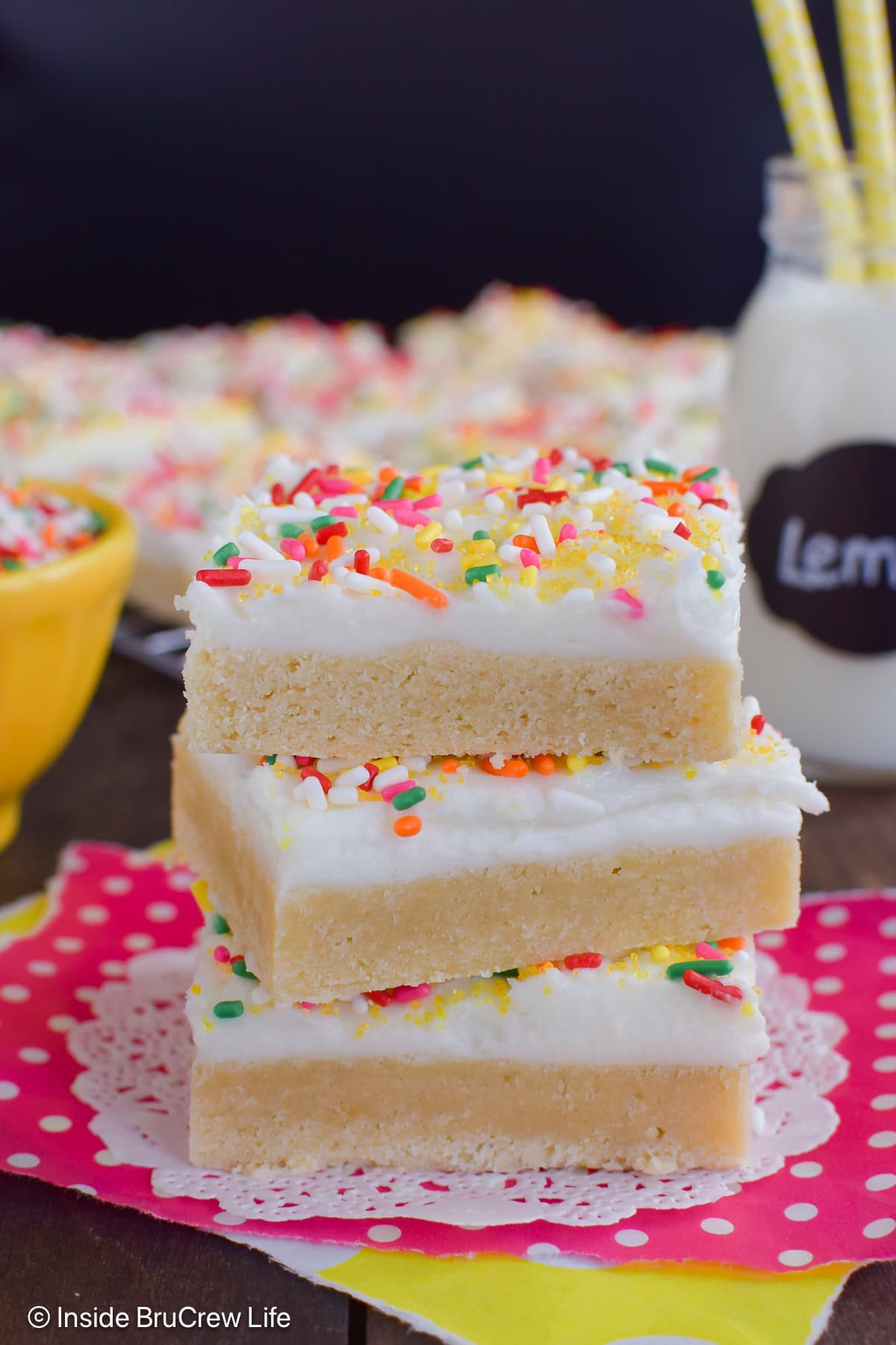 Three frosted lemon sugar cookie bars stacked on top of each other on a pink paper.