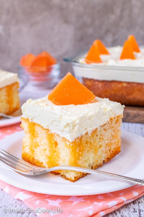 A square of vanilla cake with orange Jello stripes topped with vanilla pudding frosting and an orange slice