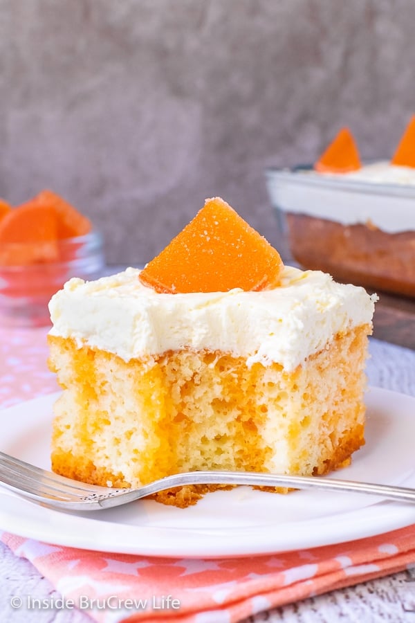 A white plate with a piece of orange creamsicle cake topped with a vanilla pudding frosting and an orange slice on it