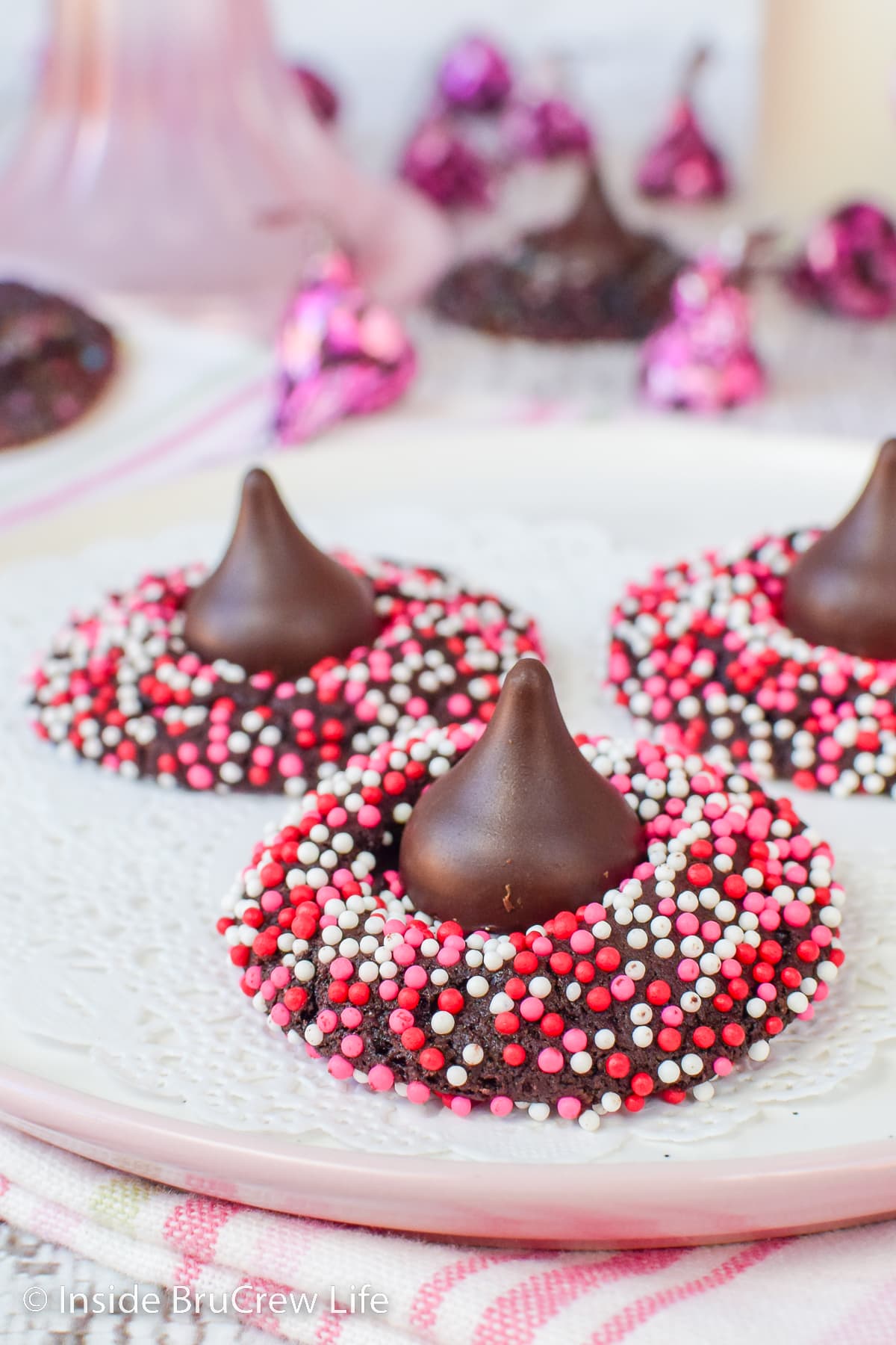 A white plate with three chocolate sprinkles cookies topped with Hershey kisses.