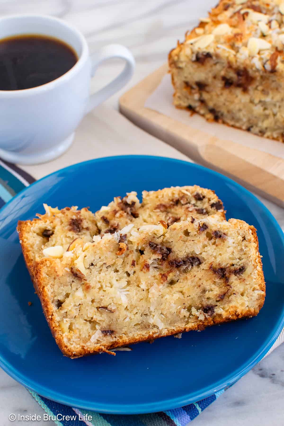 Two slices of almond joy banana bread on a blue plate with the loaf behind it.