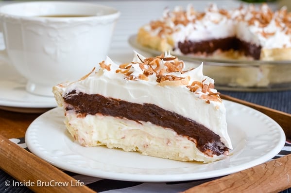 A white plate in a wood tray with a slice of coconut chocolate pie with cool whip and toasted coconut on it and the pie plate behind it