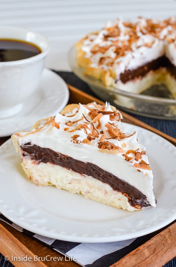 A white plate with a slice of coconut chocolate pie topped with toasted coconut on top and more pie behind it
