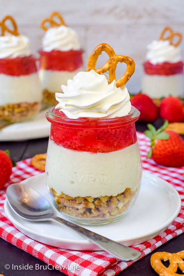 A jar of strawberry pretzel salad topped with Cool Whip and a pretzel on a white plate. More parfaits are behind it.