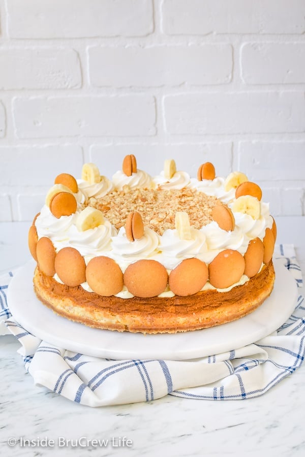 A full sized banana cheesecake topped with banana pudding on a white cake plate.