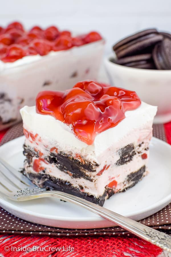 A white plate on a brown towel with a square of cherry oreo icebox cake on it and white bowl full of Oreos behind it