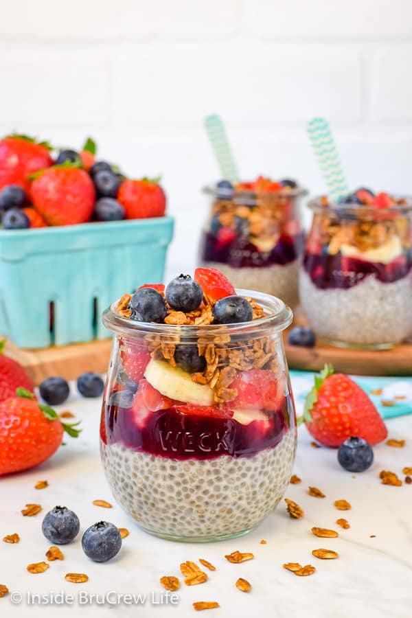 A clear jar filled with blackberry chia pudding, fruit preserves, fresh fruit, and granola and more jars behind it.