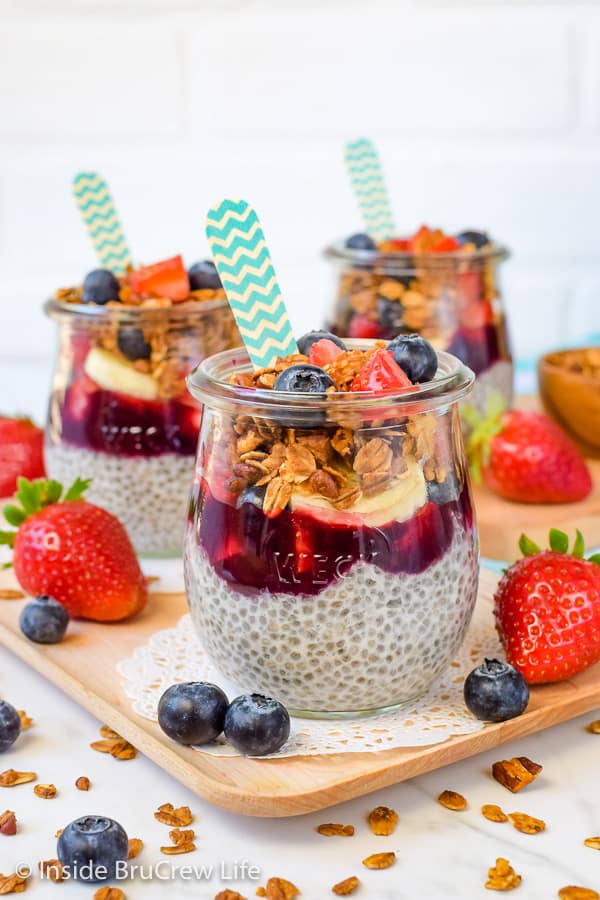 Three jars filled with chia pudding, fruit preserves, granola, and fruit with a spoon in each one.