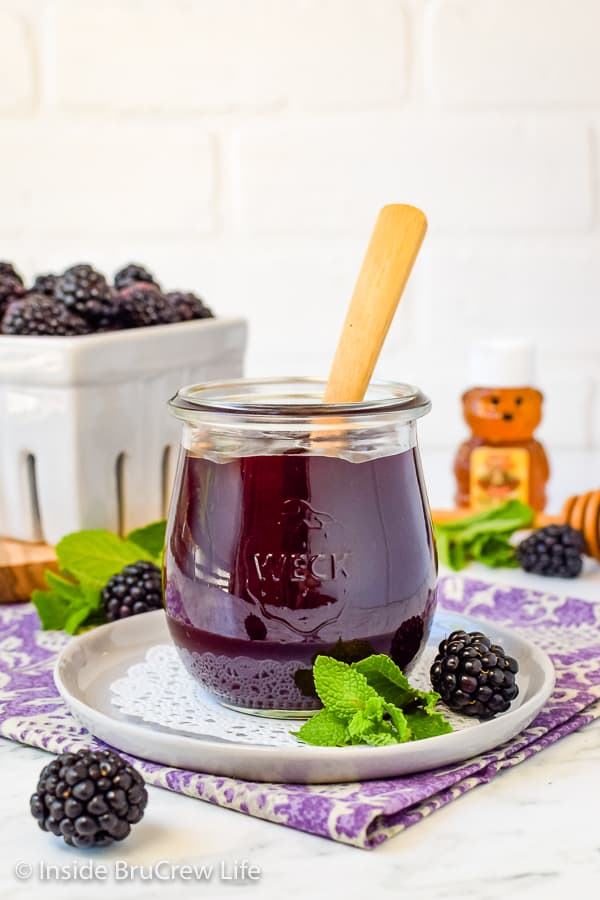 A white plate with a clear jar filled with blackberry preserves on it.