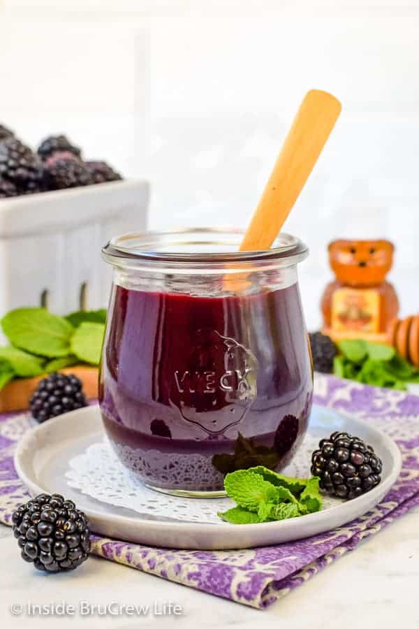 A jar of blackberry preserves on a white plate with a box of blackberries behind it.